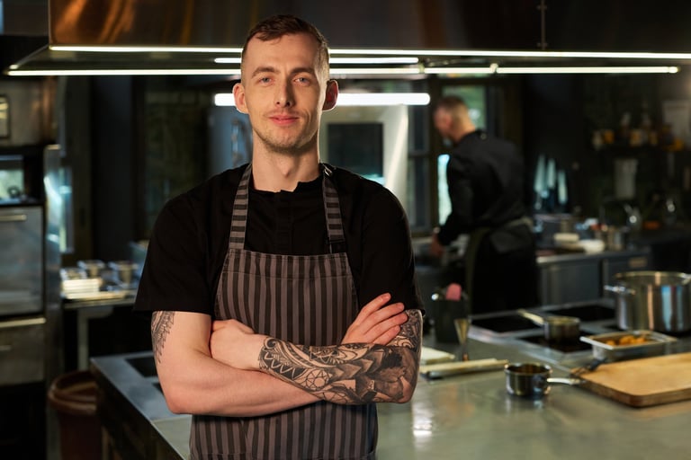 Portrait of young adult Caucasian man chef with tattoos standing in professional kitchen crossing arms and smiling confidently while another chef working in background