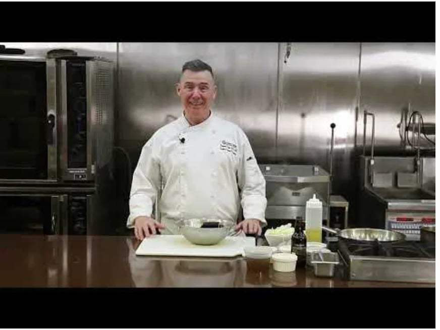 Male chef in white chefs coat standing at a counter with fresh ingredients in a professional kitchen with stainless steel equipment.