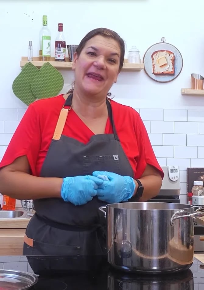 Woman in red shirt and black apron wearing blue gloves standing in kitchen with cooking pot