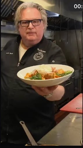 Chef wearing glasses and black jacket holds a white bowl of pasta with vegetables in a kitchen