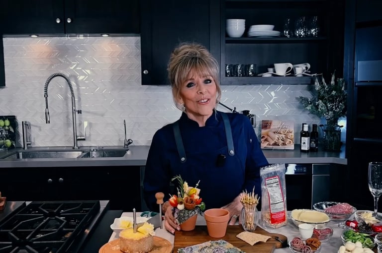 A woman with short blonde hair smiles at the camera in a modern kitchen while standing at a countertop with food and ingredients displayed in front of her.