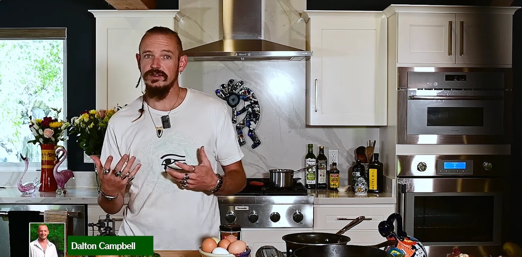 Man in white t-shirt gesturing while speaking in a modern kitchen with white cabinets and stainless steel appliances