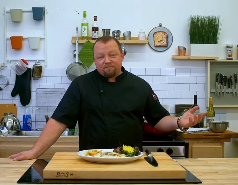 Man in black shirt standing at kitchen counter with plated food, gesturing while speaking in front of white subway tile backsplash