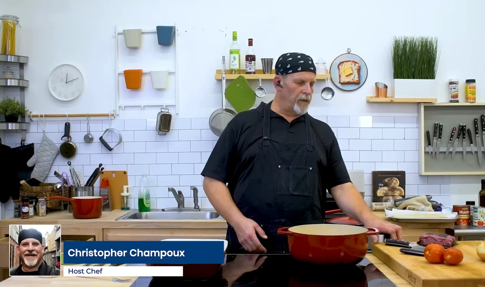 Chef Christopher Champoux in a professional kitchen with organized cookware and ingredients on white walls and shelves behind him.