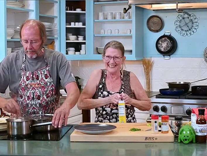 Two people cooking together in a bright kitchen, with the man stirring a pot and the woman smiling at the counter with ingredients and kitchen utensils visible.