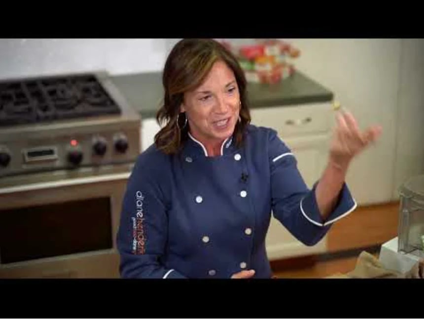 Woman in navy chef coat with white trim gesturing while speaking in a professional kitchen setting