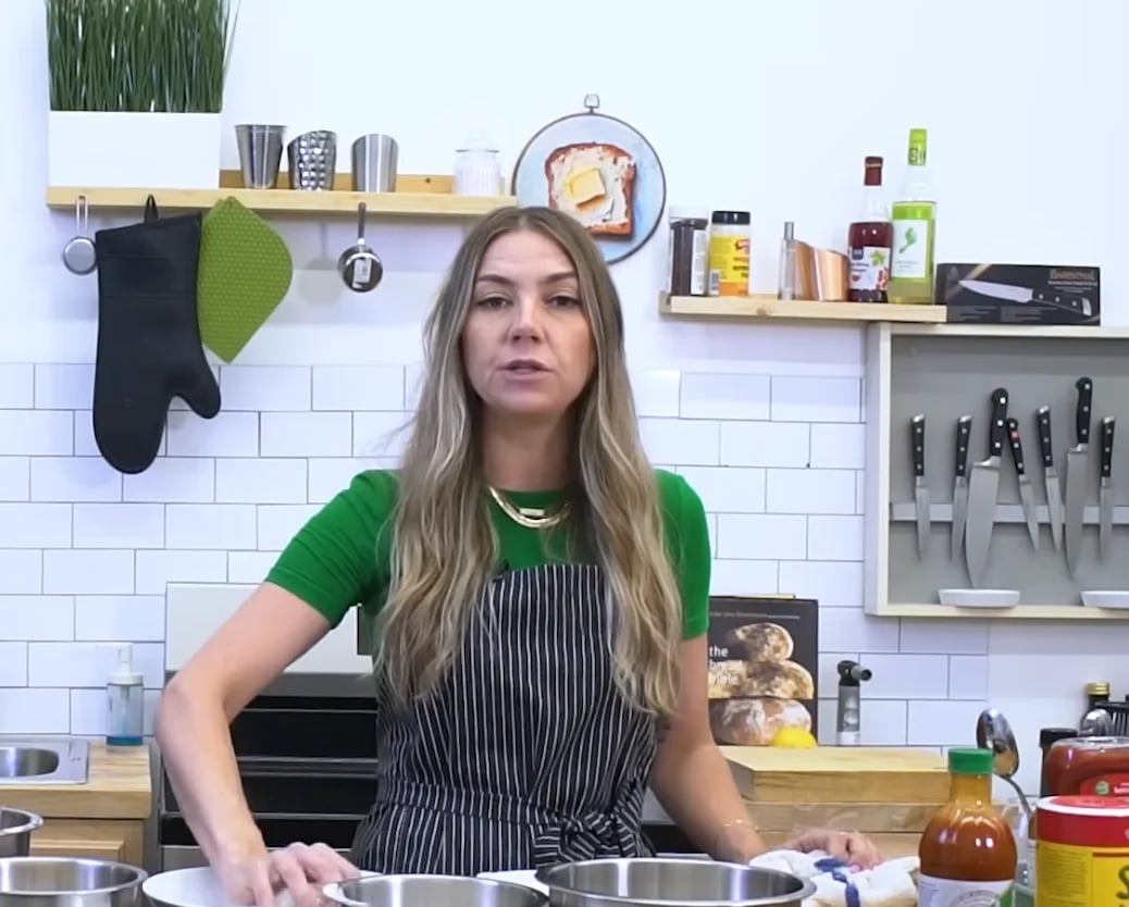 Woman in green shirt and striped apron standing in a modern kitchen with white subway tile backsplash and wooden shelves