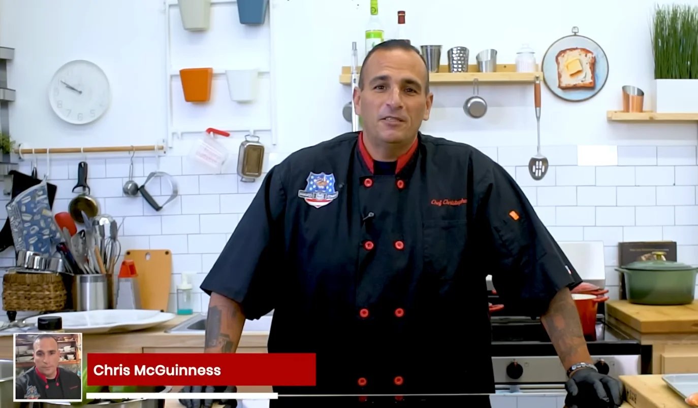 Chef Chris McGuinness in black chefs coat standing in kitchen with white subway tile backsplash and shelving